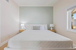 Ein ordentlich bezogenes Bett mit weißer Steppdecke, zwei Nachttischlampen und einem Fenster zur rechten Seite.