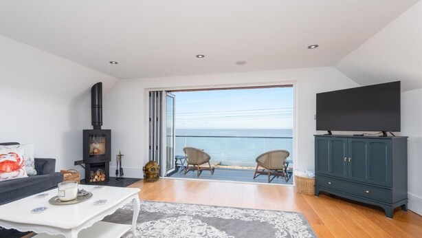 A modern living room with a large TV, a fireplace, a balcony with a view of the sea, and a cozy seating area. - Luxury Sea View House | Whitstable Beachfront (Seasalter)