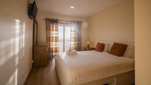 A hotel room with a bed, a TV mounted on the wall, a window with curtains, and a nightstand with a lamp. - Best Houses 64 - Sunset Beach (Baleal)