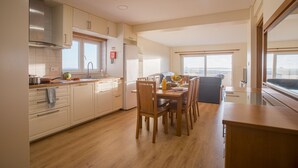 A modern kitchen with wooden cabinets, a dining table with chairs, and a large window providing a view. - Best Houses 64 - Sunset Beach (Baleal)