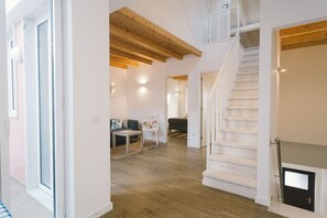 Ein großzügiger Innenraum mit einer Holztreppe, einer Sitzecke inklusive Sofa und kleinem Tisch, sowie ein heller, gut beleuchteter Raum mit großem Fenster.