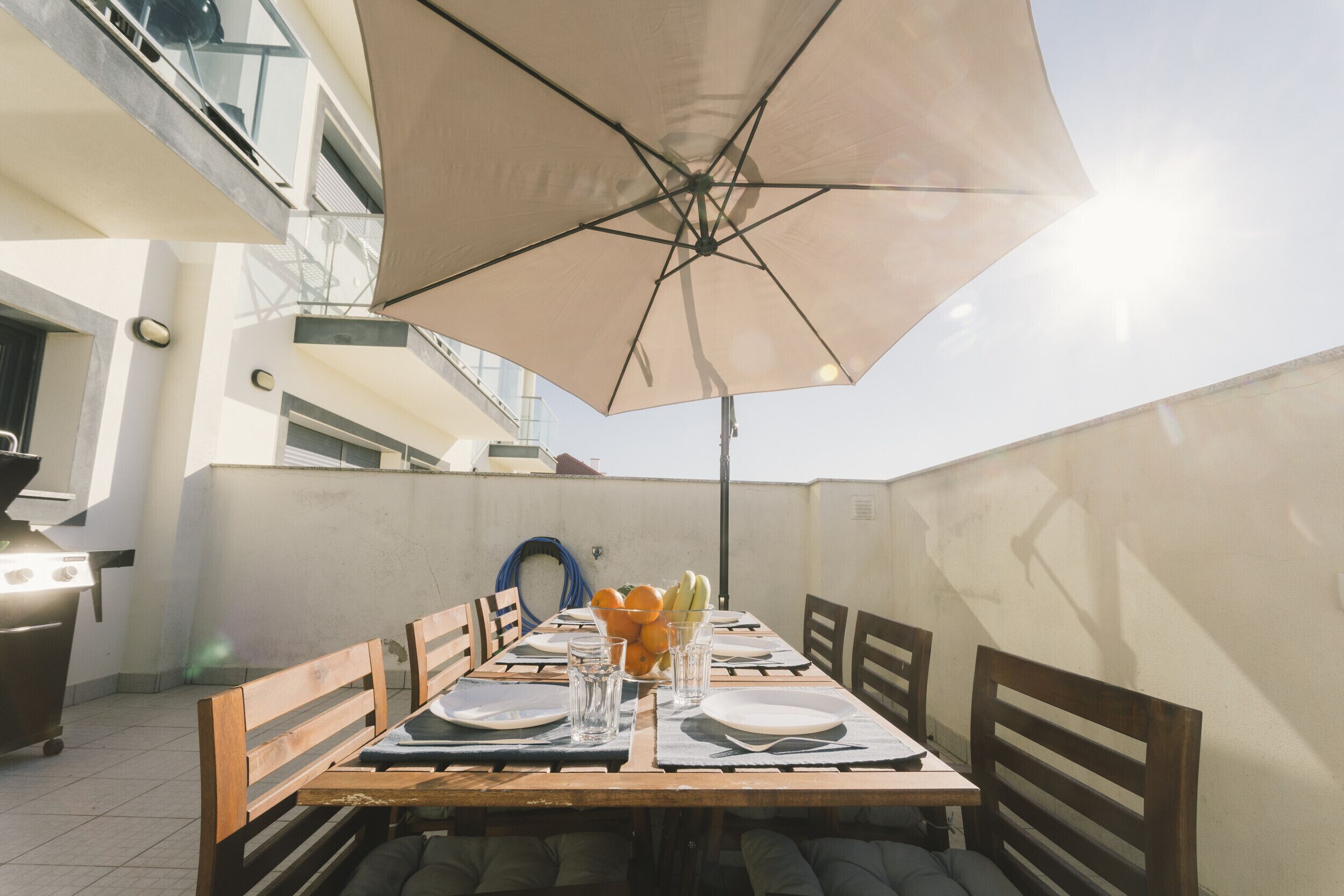 Een buiten eethoek met een tafel voor vier personen, een parasol en een barbecue op de achtergrond.