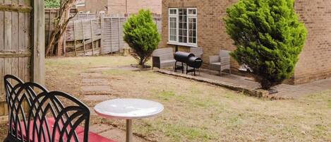 A patio with a metal chair and table, a barbecue grill, and a brick building in the background.