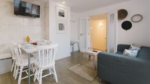Un espace de vie moderne comprenant un coin repas, un téléviseur à écran plat fixé au mur, un canapé gris et une petite table avec une télécommande.