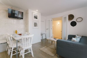 Ein zeitgemäßer Wohnbereich mit Essplatz, einem flachbildfernseher an der Wand, einem grauen Sofa und einem kleinen Tisch mit Fernbedienung.