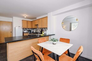 A modern kitchen with wooden cabinets, a black countertop, and a white dining table with orange chairs. - Stylish 2 BDR APT Free Parking Liverpool (Liverpool)