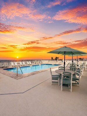 Uma área à beira da piscina com espreguiçadeiras e uma mesa, uma piscina e vista para o pôr do sol.