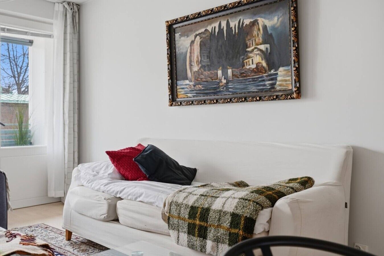 Un canapé blanc recouvert d’une couverture à carreaux, un tableau accroché au mur et une fenêtre avec des rideaux.
