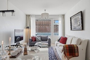 Una sala de estar moderna con un sofá, una mesa de comedor para dos personas, un televisor y un gran ventanal con cortinas.