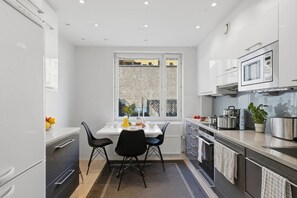 Una cocina moderna con muebles blancos, una mesa de comedor con dos sillas, un ventanal con vistas a edificios y electrodomésticos de acero inoxidable.