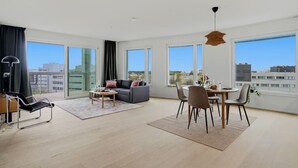 Amplia sala de estar con sofá, mesa de centro, mesa de comedor y sillas, ventanas grandes con vistas a la ciudad y una lámpara colgante moderna.
