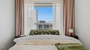 Un dormitorio con cama, almohadas, una mesita de noche, lámpara y ventana con cortinas.