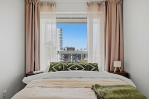 Ein Schlafzimmer mit Bett, Kissen, Nachttisch, Lampe und einem Fenster mit Vorhängen.