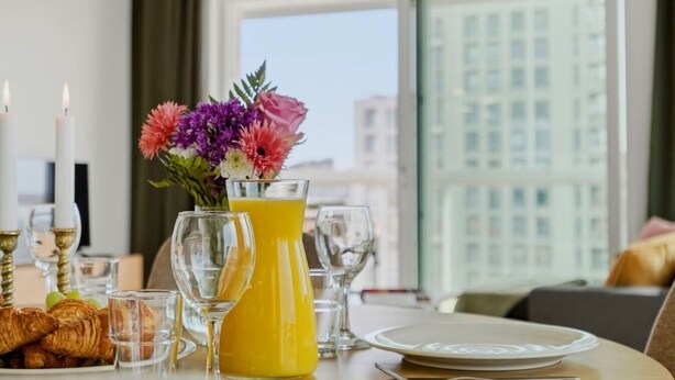 Una mesa de comedor con un jarrón de flores, una jarra de jugo de naranja y cruasanes.