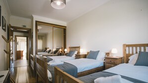 Habitación de hotel con tres camas, cabeceros de madera, mesitas de noche con lámparas, un espejo grande y suelo de madera.