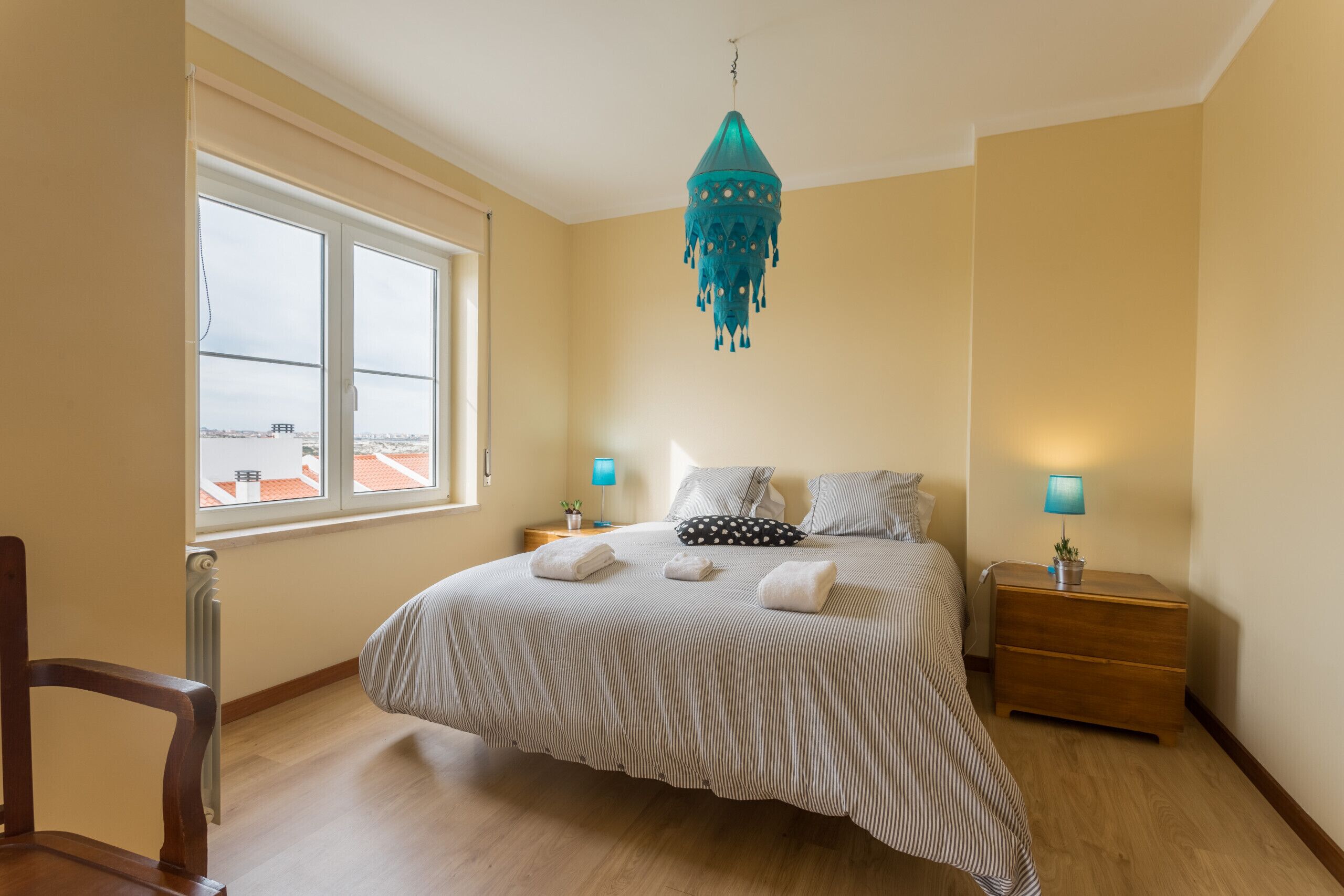 A bedroom with a large window, a bed with striped bedding, a teal chandelier, bedside lamps, and a wooden nightstand.