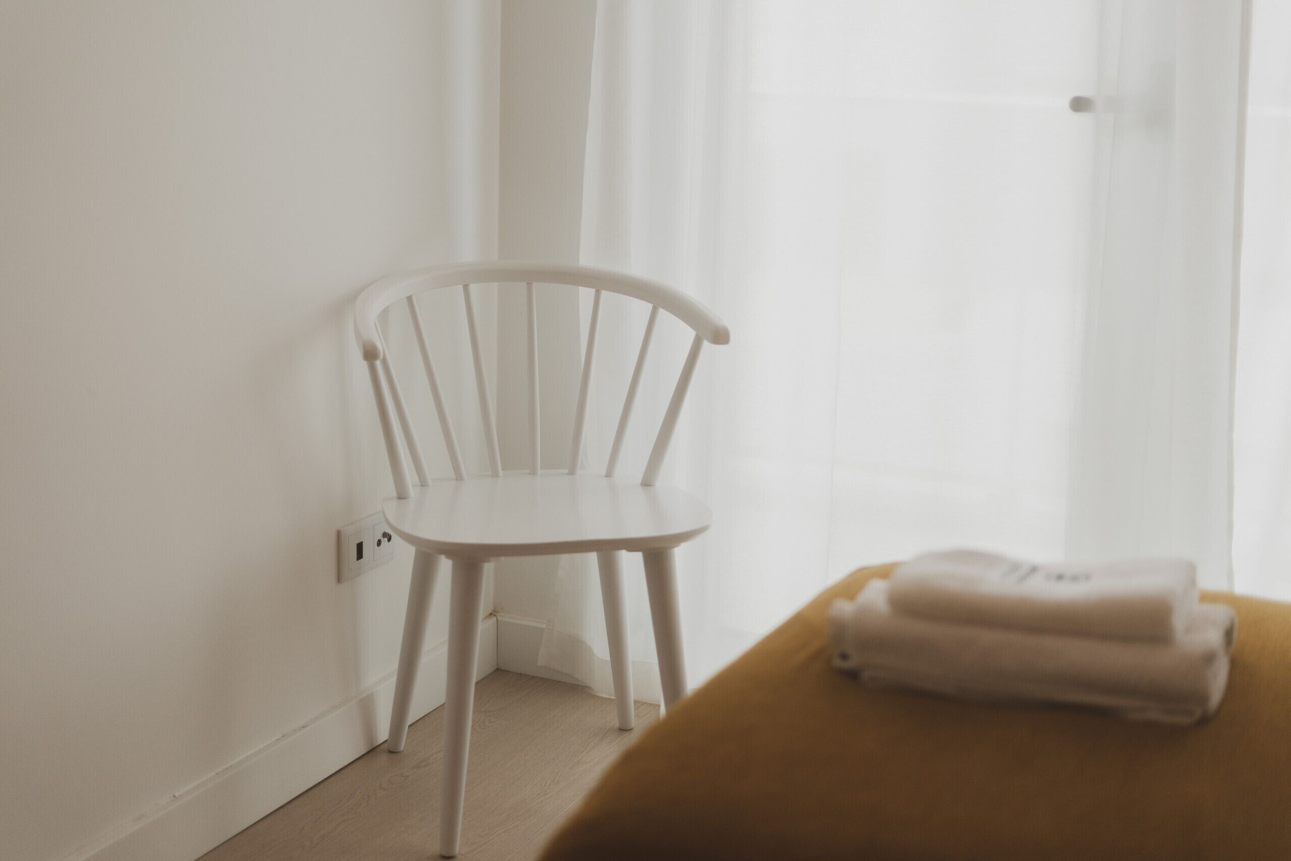 A white chair by a window with sheer curtains, a wooden table with folded towels, and a light-colored floor.