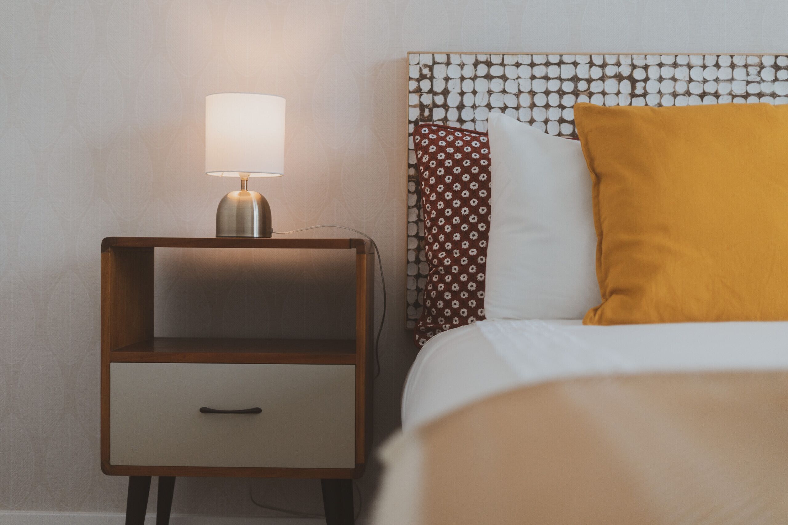 A neatly made bed with a patterned headboard, a bedside lamp, and a wooden nightstand with a drawer.