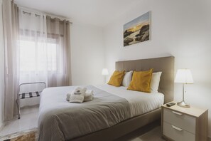 Ein ordentlich eingerichtetes Hotelzimmer mit einem Bett, Nachttischlampen, einem Beistelltisch, einem Fenster mit Vorhängen und einem Bild an der Wand.