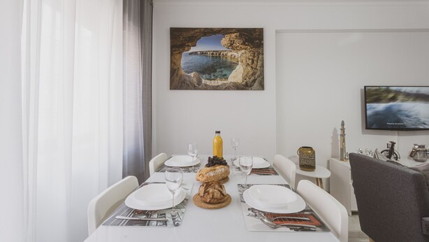 Een eetruimte met een tafel gedekt voor twee personen, een schilderij van een kustlandschap en een televisie aan de muur.