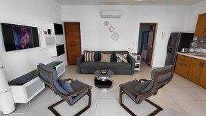 Een moderne woonkamer met een grijze bank, twee fauteuils, een salontafel, een aan de muur bevestigde televisie en een keuken met kasten en apparatuur.