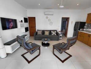 A modern living room with a grey sofa, two armchairs, a coffee table, a TV mounted on the wall, and a kitchen area with cabinets and appliances. - Charming 1-bedroom apartment with AC in Las Terrenas - LAS BAELLENAS (Las Terrenas)