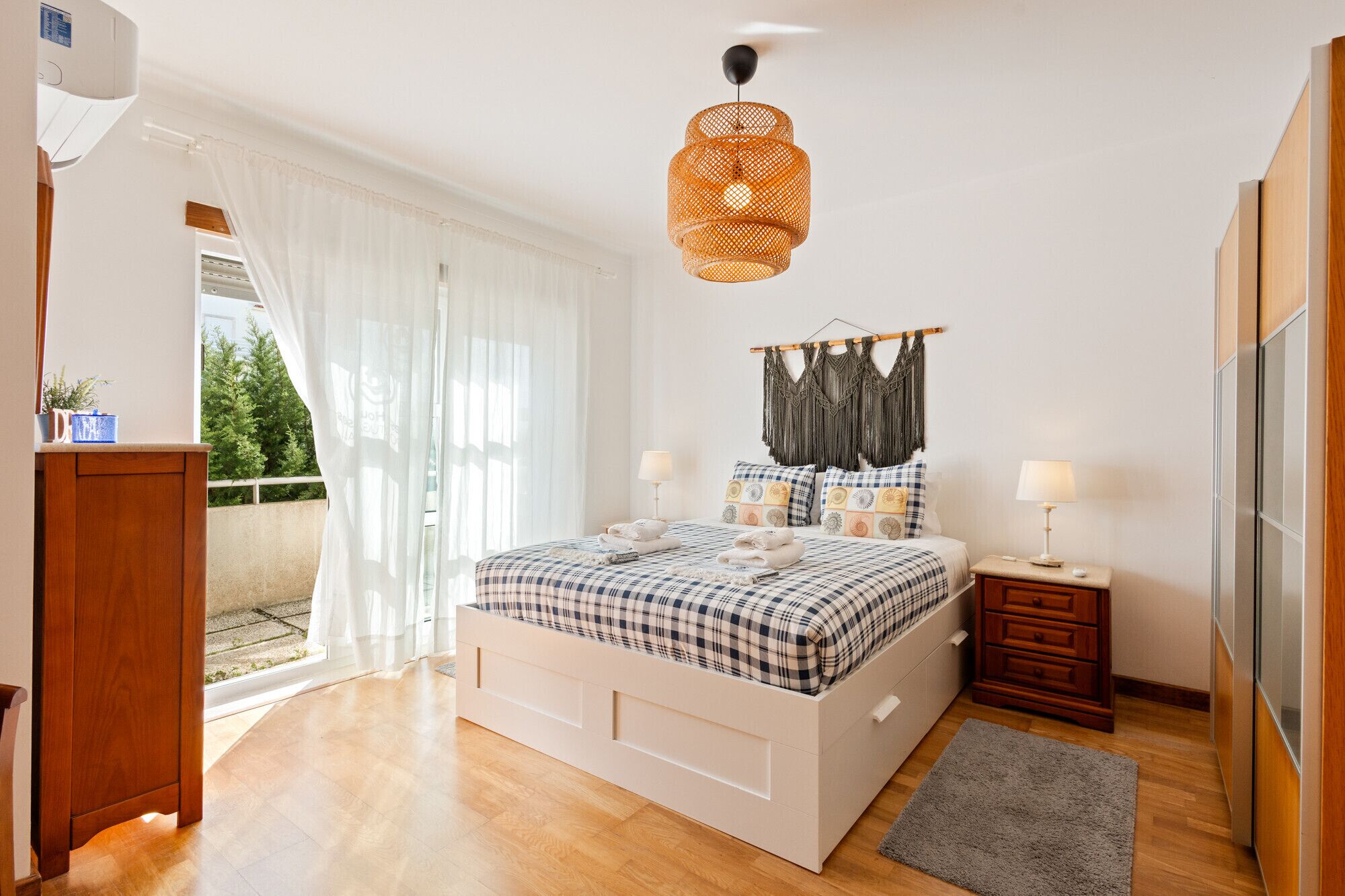 A bedroom with a bed, bedside tables, a hanging macrame decoration, and a view of greenery outside.