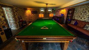 Uma mesa de bilhar numa sala com paredes de pedra, um ventilador de teto e um espaço de bar com estantes de livros.