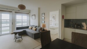Sala de estar con sofá, una mesa redonda, lámpara y sillón. Se observa una zona de cocina con armarios y encimera.