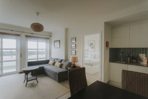 A living room with a sofa, a round table, a lamp, and a chair. There is a kitchen area with cabinets and a countertop. - Best Houses 40 - You me and the sea (Baleal)