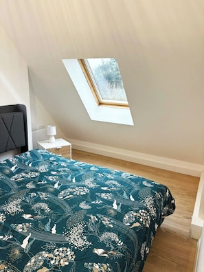 Ein Schlafzimmer mit Dachfenster, einem Bett mit floralem Überwurf, einem Nachttisch mit Lampe und einem dunklen Kopfteil.