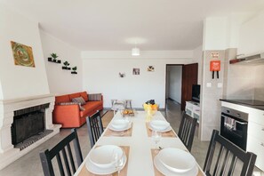 Un comedor con una mesa blanca dispuesta para cuatro, chimenea, sofá y zona de cocina con estufa.