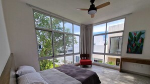 Ein Schlafzimmer mit einem Bett, einem Sessel, einem Deckenventilator, großen Fenstern und einem Bild an der Wand.