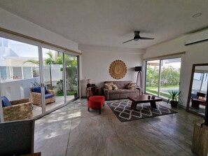 A spacious living area with a sofa, a red chair, a coffee table, and a floor lamp. There is a large window with a view of greenery outside. - House with Private Pool15 min Apt 20 MIn Hotel Zone (Cancún)