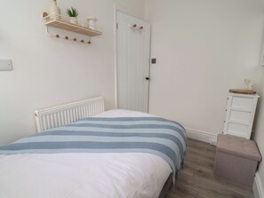 A bedroom with a bed, a radiator, a white door, a small white dresser, and a shelf with decorative items. - Driftwood (Rhyl)