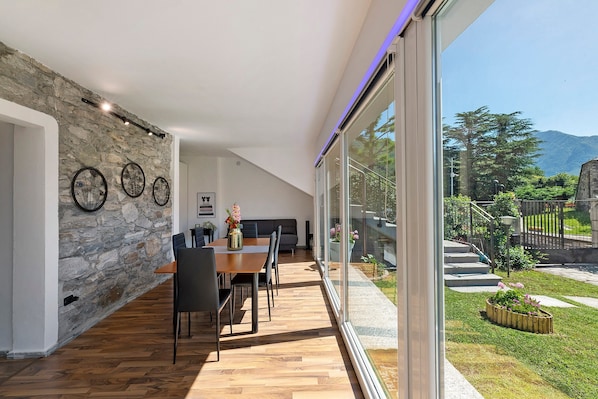 Um interior moderno com parede de pedra, piso de madeira, área de jantar e uma grande janela de vidro que oferece vista para um jardim e montanhas.
