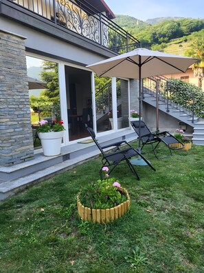 A modern house with a stone facade, a balcony with metal railings, a covered patio with chairs, and a view of a hillside.