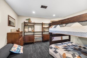 Un lit superposé avec une courtepointe aux motifs géométriques, une commode en bois, une bibliothèque et un sol recouvert d’un tapis gris.