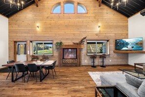 Ein geräumiges Zimmer mit Holzbalkendecke, Kamin, Esstisch mit Stühlen und einem großen, an der Wand befestigten Fernseher.