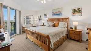 A bedroom with a bed, nightstands, a desk, a chair, a ceiling fan, and a view through a sliding door. - Oceanwalk 15-403 (New Smyrna Beach)