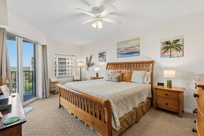 A bedroom with a bed, nightstands, a desk, a chair, a ceiling fan, and a view through a sliding door. - Oceanwalk 15-403 (New Smyrna Beach)
