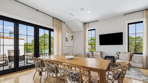 Un comedor amplio con una larga mesa de madera, sillas de mimbre con cojines y grandes ventanales que ofrecen vistas al exterior.