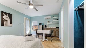 Un dormitorio con cama, ventilador de techo, mesa de comedor con sillas, zona de cocina con armarios y refrigerador, y aire acondicionado instalado en la pared.