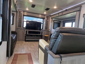 A compact RV interior with a seating area, a television, and a window providing a view.