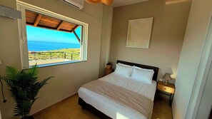 Una camera da letto con un letto, un comodino, una lampada, una pianta in vaso, una finestra vista mare e una ventola a soffitto.