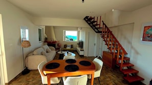 Ein Wohnzimmer mit einer Couch, einem Esstisch mit Platzsets, einer Treppe und Blick durch ein Fenster.