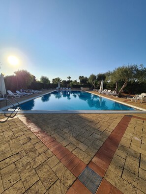 Ein Pool mit Liegestühlen und Sonnenschirmen, umgeben von Bäumen und blauem Himmel.