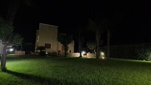 Een nachtzicht van een huis met een verlicht gazon, een stenen muur en enkele bomen.