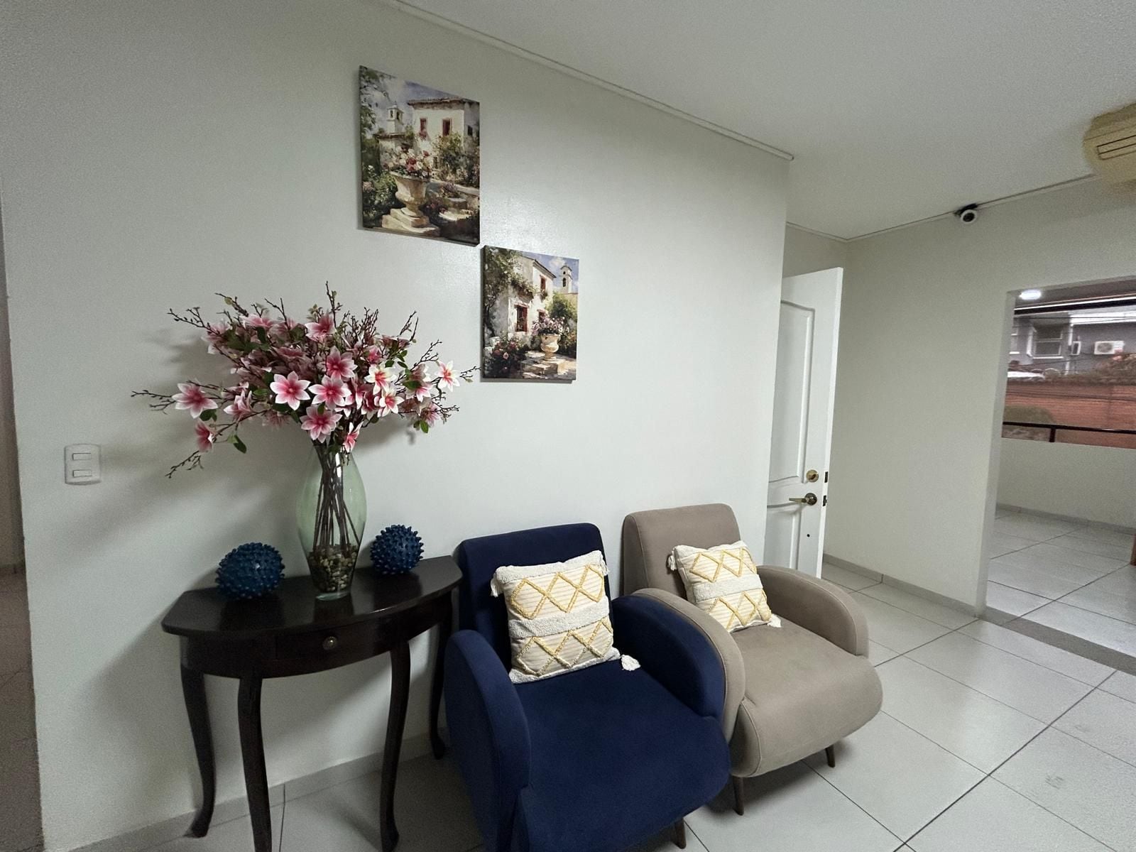 A living room with a sofa, armchairs, a small table with a vase of flowers, and two wall-mounted pictures.