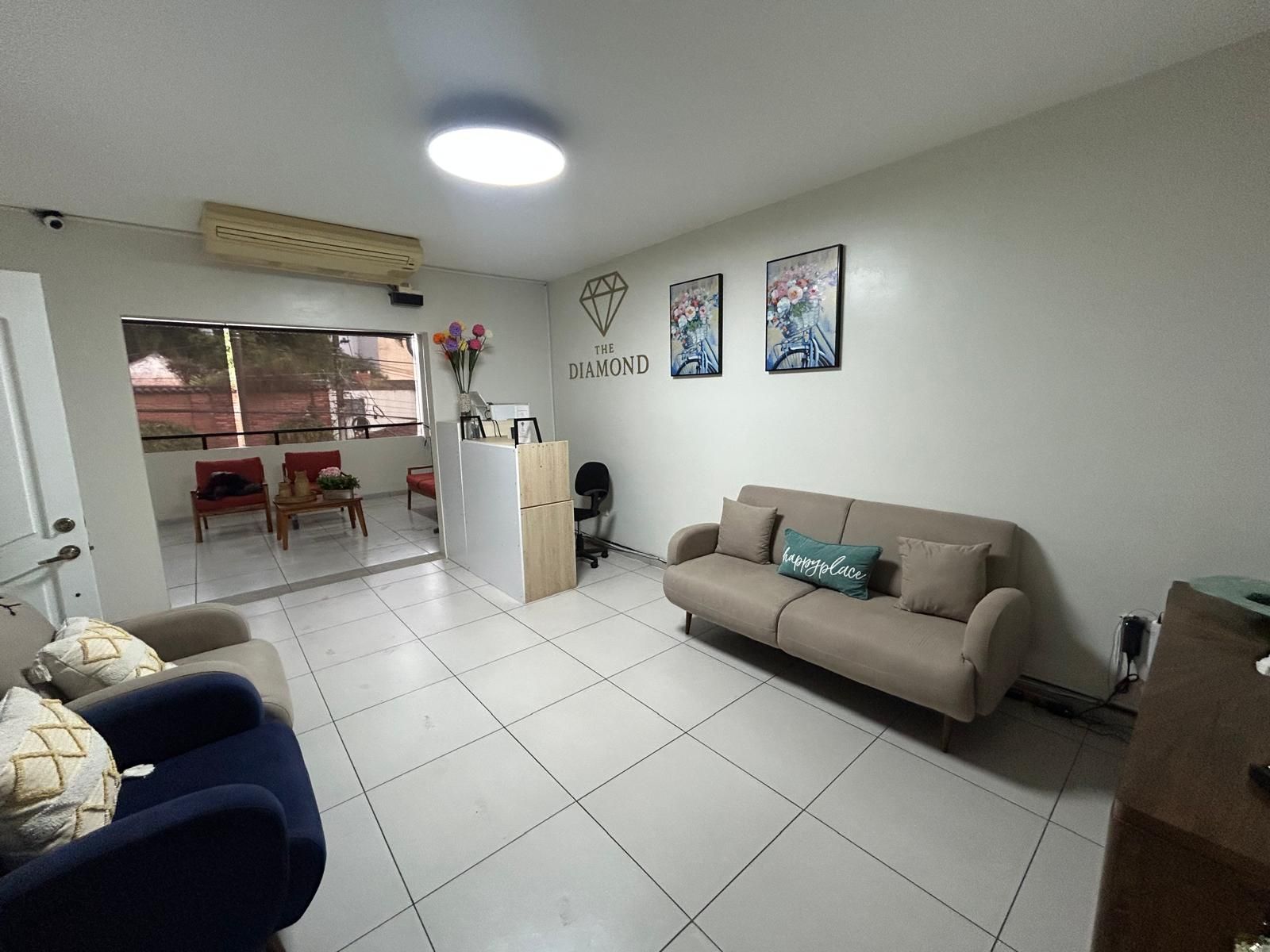 A living room with a beige sofa, a coffee table, and a red chair. There is a small desk with a computer and a vase with flowers. The room has a tiled floor and a window with a view outside.